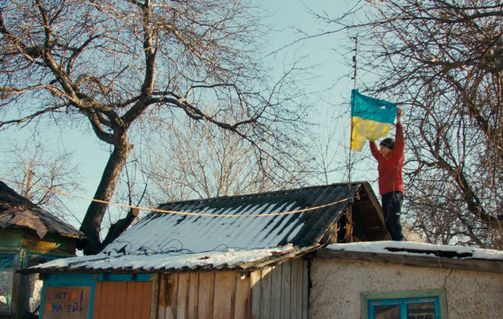 Eine Person steht auf dem Dach eines Schuppens und hält eine blau-gelbe Flagge hoch. Es ist die ukrainische Flagge.