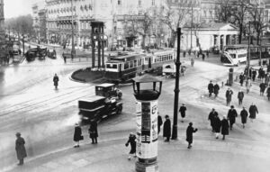 Schwarz/Weiß: Die Kreuzung einer Großstadt in den 1920er Jahren mit Passanten, Autos und einer Straßenbahn sind zu sehen.