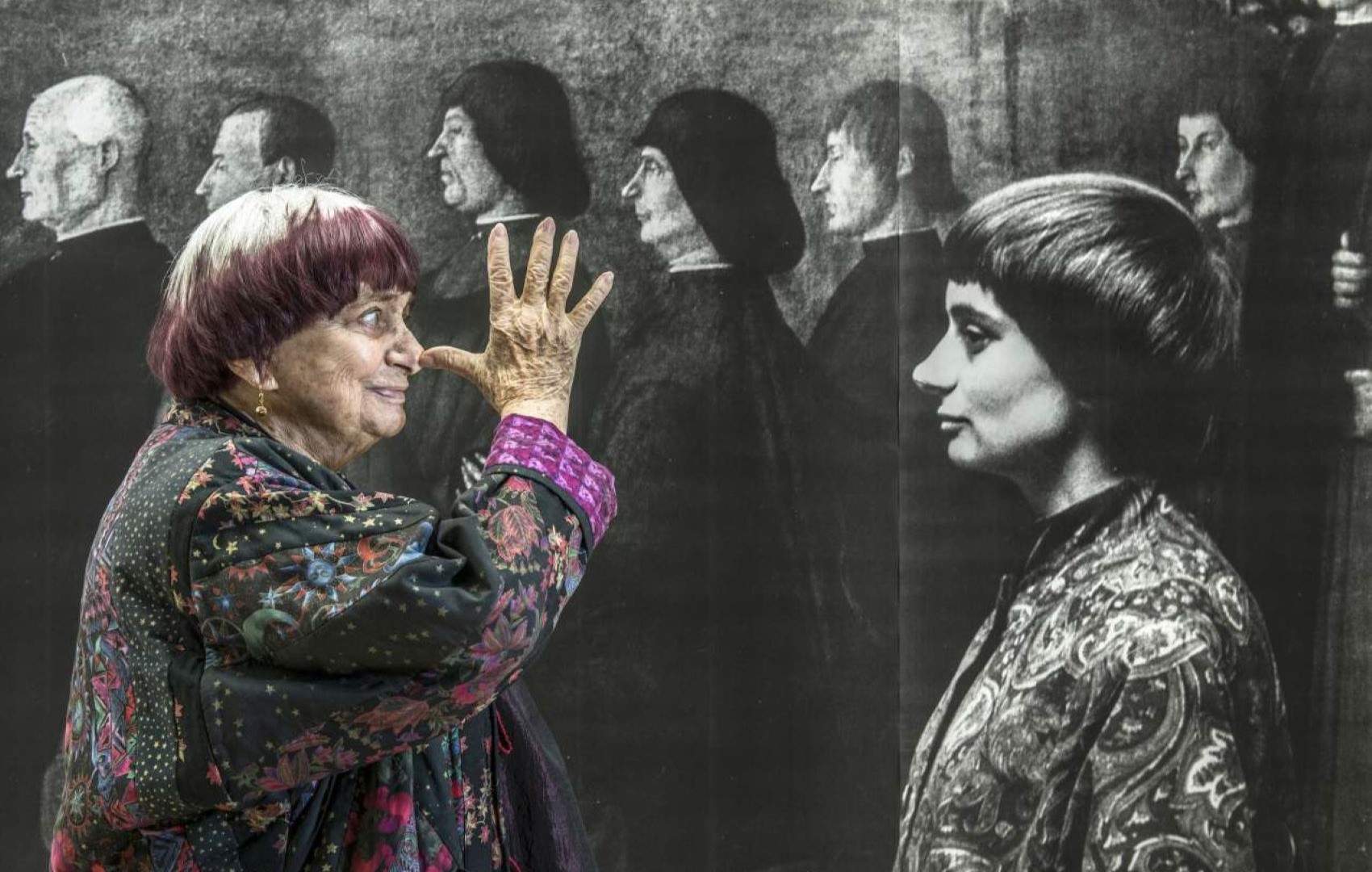 Die Regisseurin Agnes Varda blickt mit einer frechen Handgeste auf eine Wand mit Fototapete, auf der sie in jungen Jahren abgebildet ist.